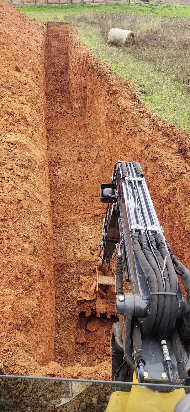 A deep trench cut through red clay with the excavator boom working in it — Little Acres Grading