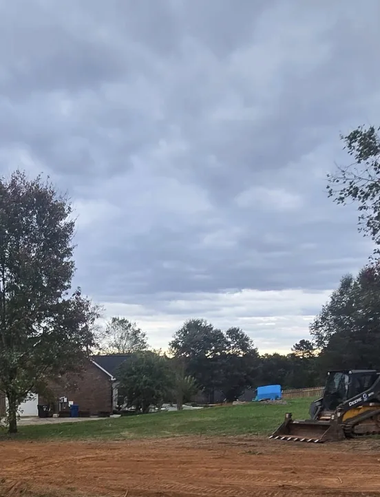 A skid steer finishing grade beside a brick home at dusk — Little Acres Grading