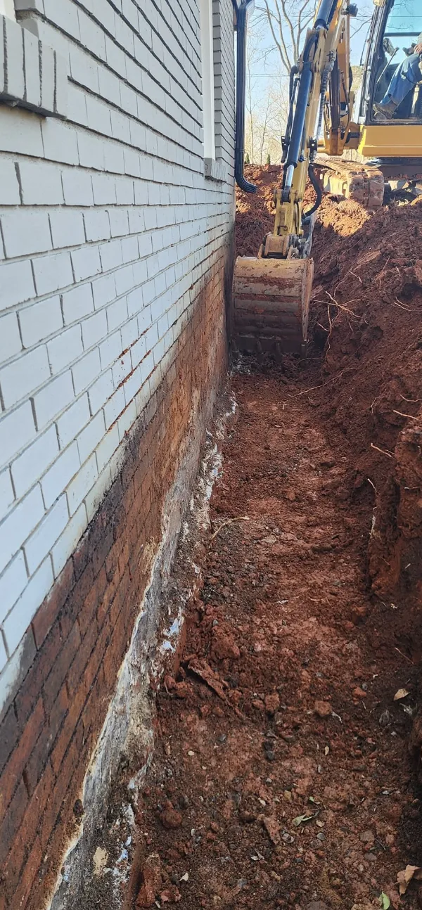 A foundation trench opened along the wall of a white brick home — Little Acres Grading