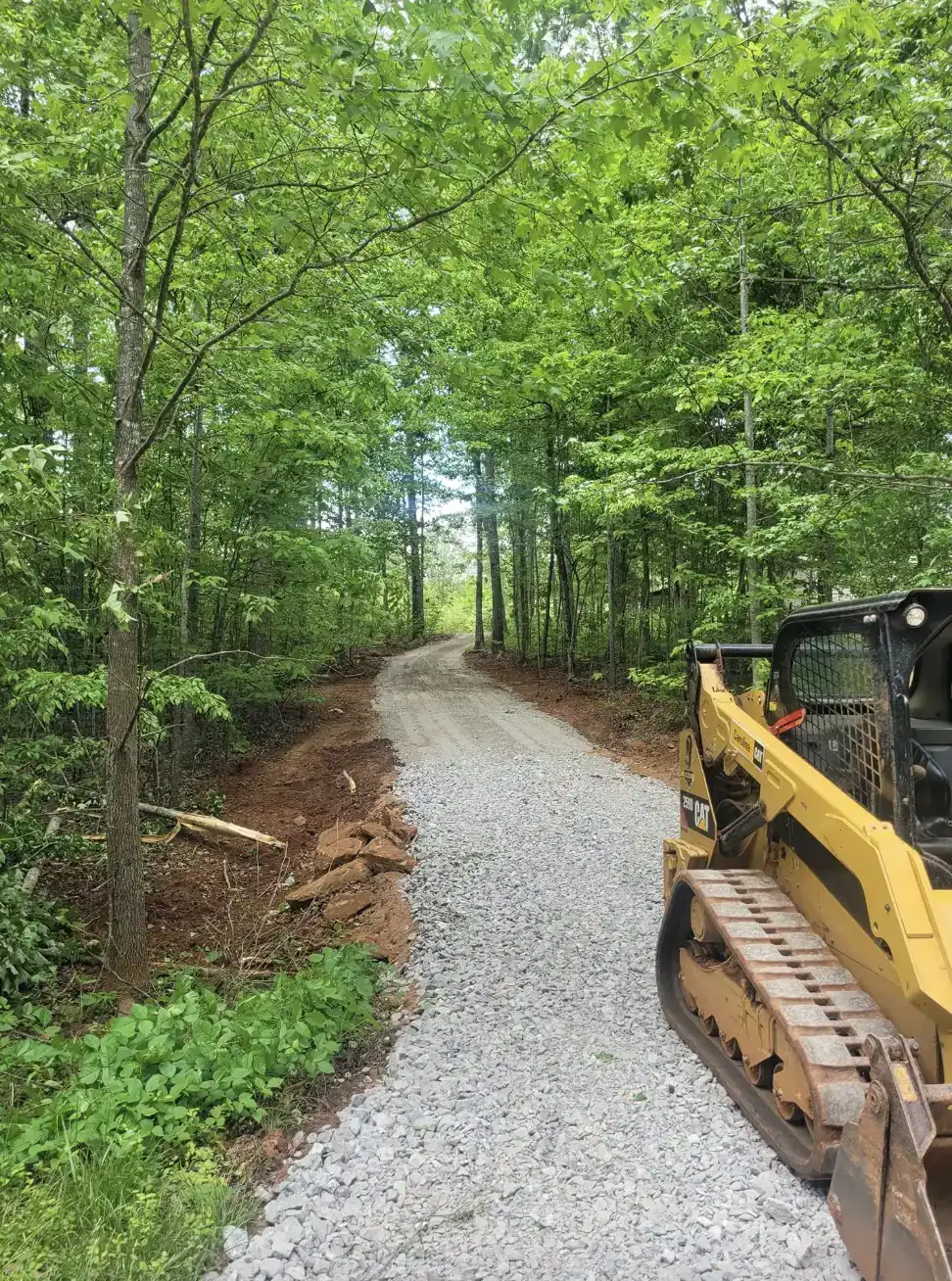 A skid steer alongside a newly cut gravel road through the woods — Little Acres Grading